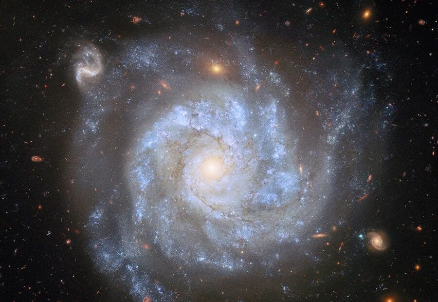 Uma visão de cima de uma galáxia espiral, mostrando seu centro brilhante, seus amplos braços espirais e o halo tênue ao redor de seu disco, além de galáxias e estrelas distantes sobre um fundo escuro. Grandes nuvens azuis de gás, salpicadas com pequenas estrelas e fios de poeira escura, giram ao redor do disco da galáxia. Algumas das galáxias ao fundo são grandes o suficiente para que seus próprios braços espirais giratórios sejam visíveis.