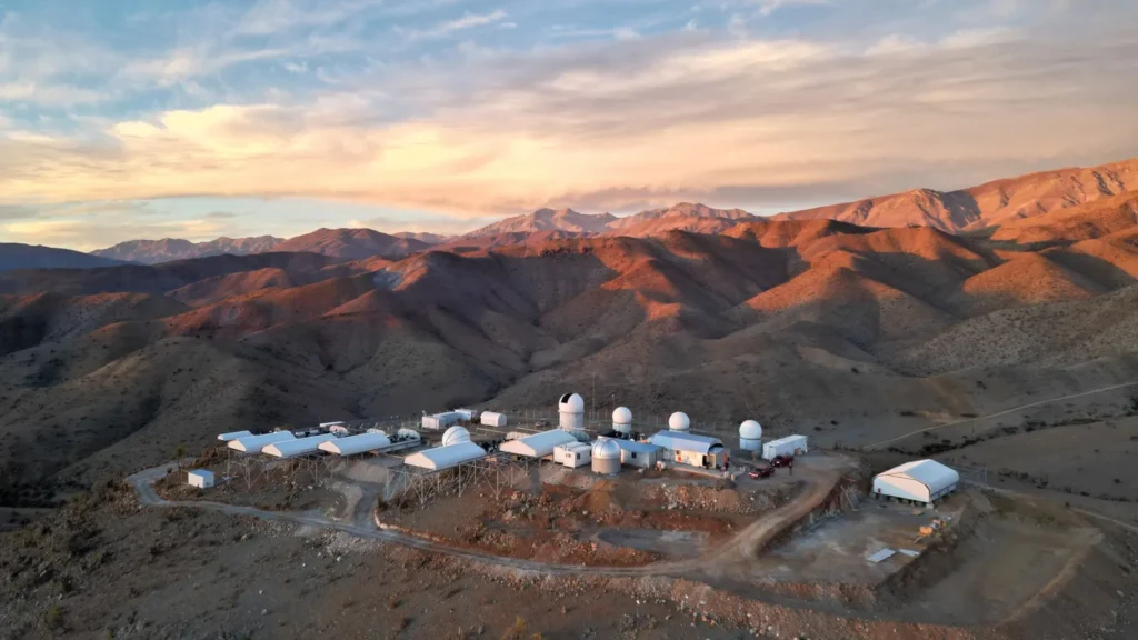 Complexo do Observatório El Sauce localizado entre cadeias de montanhas ao pôr do sol, com estruturas brancas destacando-se sob o céu alaranjado.