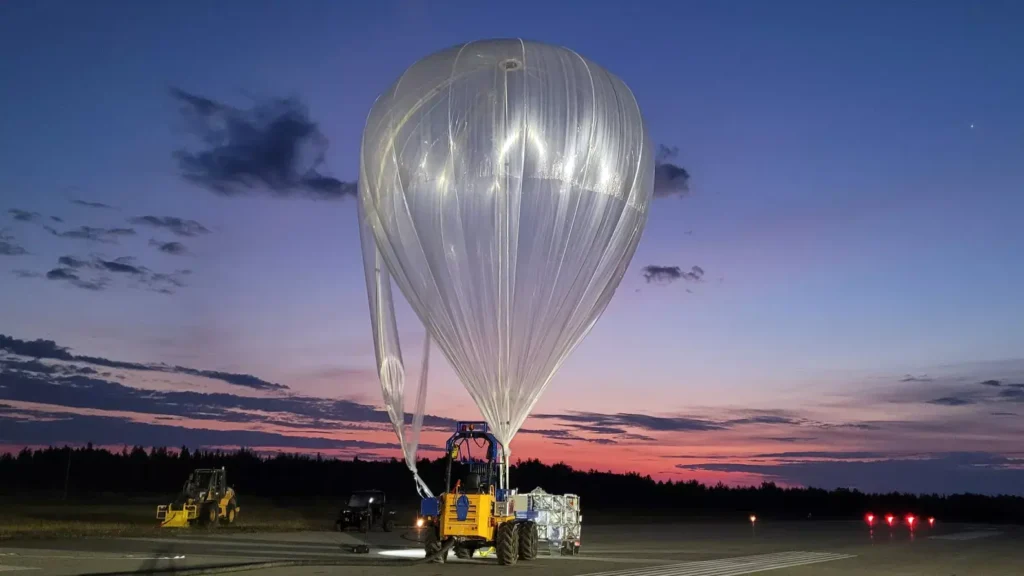 Pessoas trabalham na preparação de um balão estratosférico gigante, inflando sua estrutura branca enquanto o céu começa a ganhar tons alaranjados e rosados com o romper da aurora ao fundo.