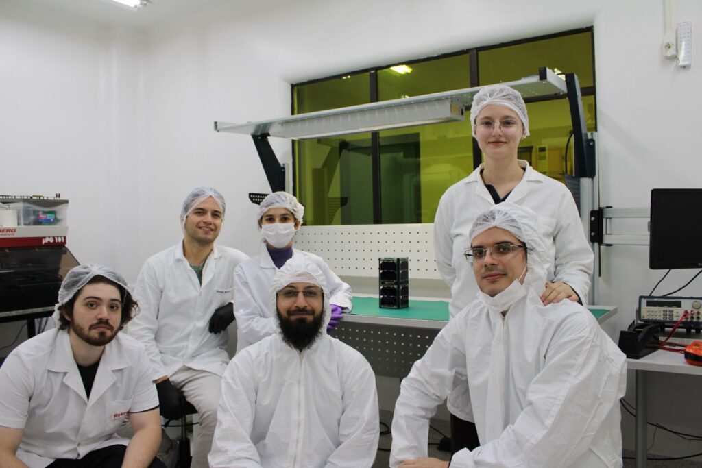 Equipe do SpaceLab posando em laboratório, com o nanosatélite ao fundo. Cientistas com equipamentos e instrumentos, em ambiente de pesquisa. Foto: Eduardo Bezerra/UFSC.