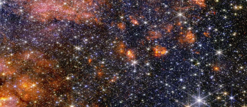 Uma visão ampla de uma região do espaço repleta de estrelas e aglomerados de nuvens alaranjadas.