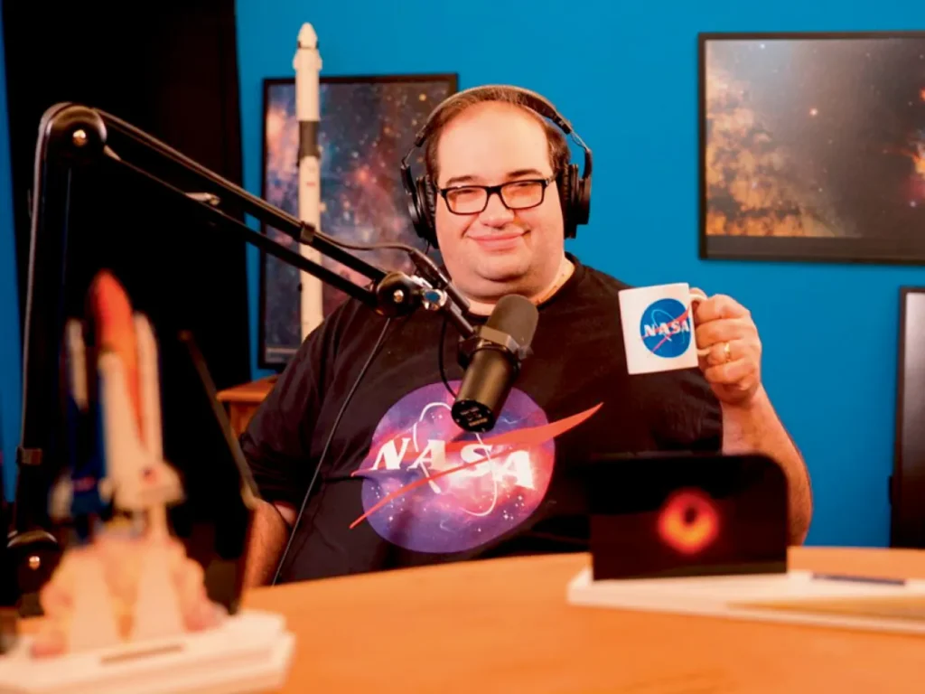 Sergio Sacani no estúdio do podcast Ciência Sem Fim, segurando uma caneca da NASA e usando uma camiseta com o logotipo da agência. Ao fundo, imagens espaciais decoram o cenário.
