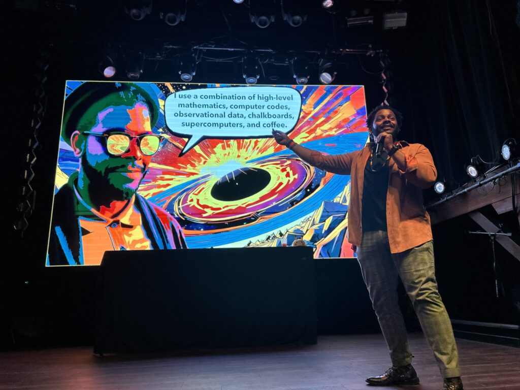 Ronald Gamble, funcionário da NASA, em primeiro plano, com uma apresentação colorida sobre um buraco negro supermassivo exibida ao fundo, destacando pesquisa e divulgação científica. Crédito: The Planetary Society