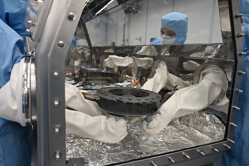 Técnicos da equipe de curadoria da missão OSIRIS-REx posicionam o mecanismo TAGSAM dentro da glovebox do laboratório, manipulando cuidadosamente o coletor de amostras do asteroide Bennu para processamento seguro.