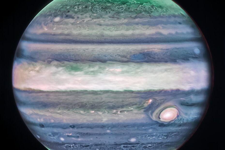 Júpiter domina o fundo preto do espaço. A imagem é uma composição em cores aprimoradas, mostrando o planeta com sua turbulenta Grande Mancha Vermelha — que aqui aparece em branco. O planeta exibe faixas horizontais onduladas em tons de turquesa neon, azul lavanda, rosa-claro e creme. Essas faixas se misturam nas bordas, como creme sendo misturado ao café. Em ambos os polos, o planeta brilha em turquesa, enquanto auroras alaranjadas intensas reluzem logo acima da superfície. Créditos: NASA, ESA, CSA, STScI