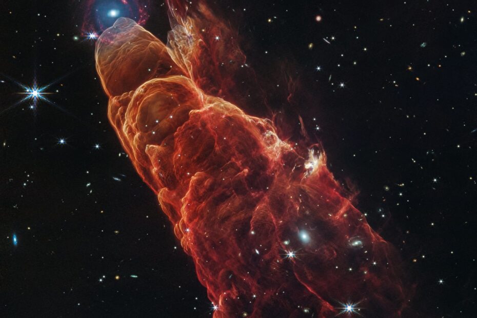 Da parte superior esquerda até a inferior direita da imagem, estende-se uma nuvem em formato de cone, em tons de laranja e vermelho, conhecida como Herbig-Haro 49/50. Essa estrutura ocupa cerca de três quartos do comprimento total da imagem. A ponta do cone, localizada no canto superior esquerdo, é translúcida e possui uma extremidade arredondada. Coincidentemente, bem próxima a essa ponta, há uma galáxia espiral ao fundo, com um centro azul brilhante que se desfaz gradualmente em braços espirais avermelhados. A estrutura em forma de cone se alarga levemente em direção à parte inferior direita, apresentando bordas arredondadas — como ondas — e detalhes intricados que lembram espuma. A nebulosa torna-se ainda mais translúcida nessa região, revelando melhor o fundo negro do espaço, pontilhado por estrelas brancas e pequenas galáxias tênues distribuídas por toda a cena.