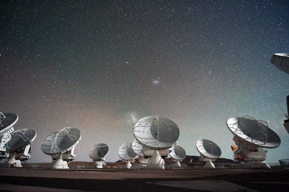 O observatório Atacama Large Millimeter/submillimeter Array (ALMA) visto à noite, com suas antenas iluminadas sob as Nuvens de Magalhães no céu do deserto do Chile.