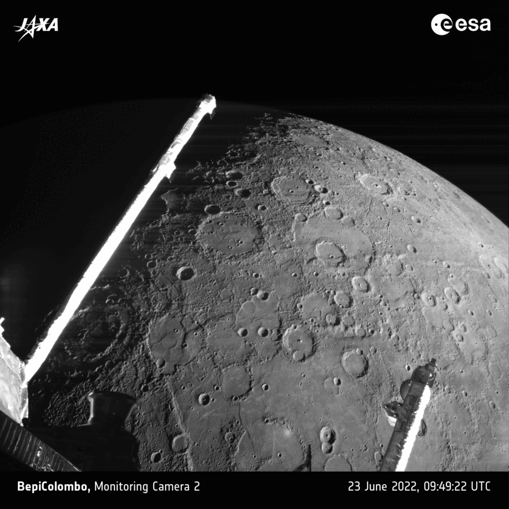 Imagem em preto e branco da superfície de Mercúrio capturada pela sonda BepiColombo durante um sobrevoo em 23 de junho de 2022. A cena mostra crateras, escarpas e terrenos de diferentes texturas, com o braço do magnetômetro da espaçonave atravessando o quadro diagonalmente.