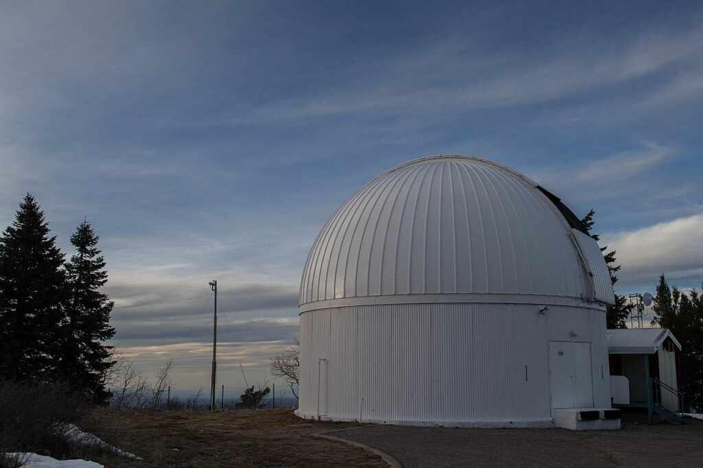 Observatório Catalina Sky Survey ao entardecer, com cúpula branca em primeiro plano e céu parcialmente nublado nas Montanhas Santa Catalina, Arizona.