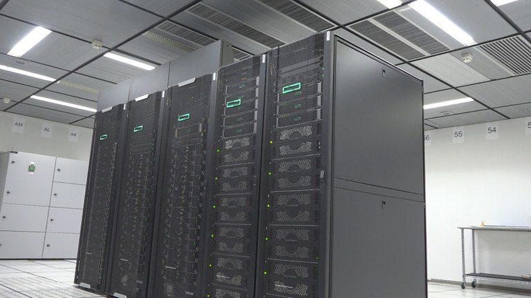 Sala do datacenter do INPE com o supercomputador Jaci. O ambiente é iluminado por luzes brancas e azuis, mostrando fileiras de racks metálicos que abrigam servidores de alto desempenho. Cabos organizados e painéis de refrigeração indicam o poder computacional dedicado a pesquisas climáticas e espaciais. Crédito: INPE