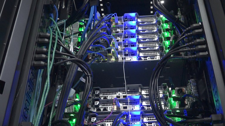 Sala do datacenter do INPE com o supercomputador Jaci. O ambiente é iluminado por luzes brancas e azuis, mostrando fileiras de racks metálicos que abrigam servidores de alto desempenho. Cabos organizados e painéis de refrigeração indicam o poder computacional dedicado a pesquisas climáticas e espaciais. Crédito: INPE