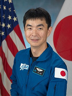 Retrato oficial do astronauta japonês Kimiya Yui em traje azul da JAXA, posando diante das bandeiras do Japão e dos Estados Unidos, em estúdio, olhando diretamente para a câmera.