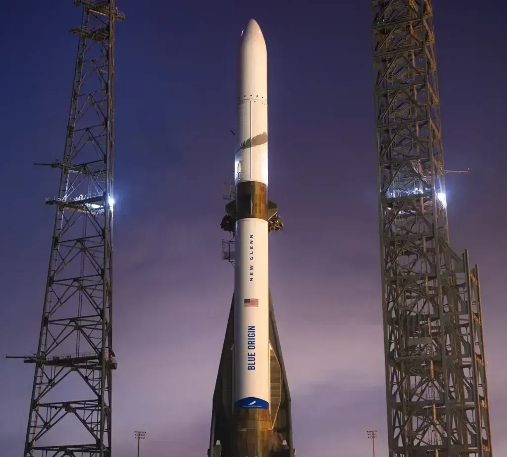 Foguete New Glenn da Blue Origin parado na plataforma de lançamento durante o entardecer, com o céu alaranjado iluminando a estrutura do veículo.