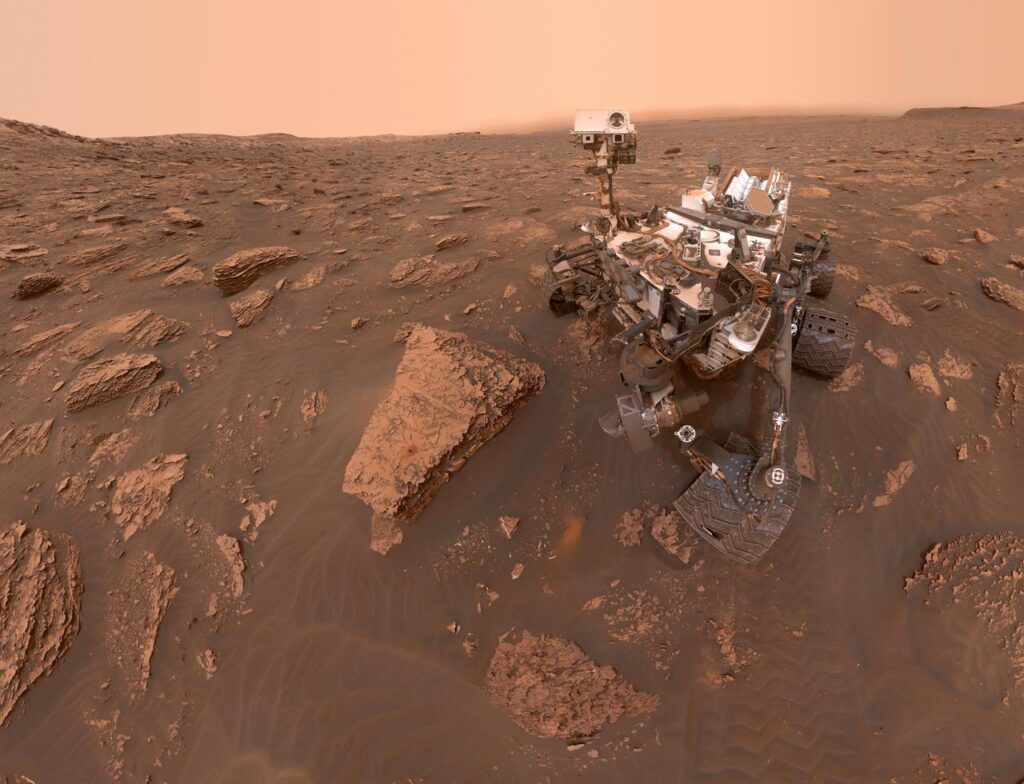 Autorretrato do rover Curiosity em Marte durante uma tempestade de poeira, com o ambiente ao redor escurecido pela baixa visibilidade no Cratera Gale. A imagem foi capturada pela câmera MAHLI no Sol 2082. Credit: NASA/JPL-Caltech/MSSS