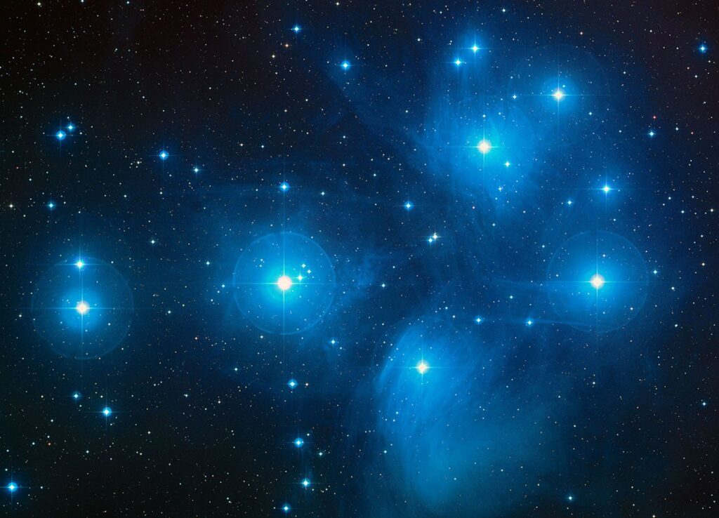 Imagem colorida das Plêiades mostrando estrelas jovens e azuladas espalhadas pela constelação de Touro, capturadas pelo telescópio Schmidt do Observatório Palomar. As estrelas brilhantes do aglomerado, a cerca de 430 anos-luz, aparecem nitidamente dispersas no céu. Crédito: NASA/ESA/AURA/Caltech.