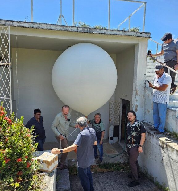 Equipe de seis pessoas realiza a preparação de um balão estratosférico branco antes do lançamento, em área aberta sob céu azul.