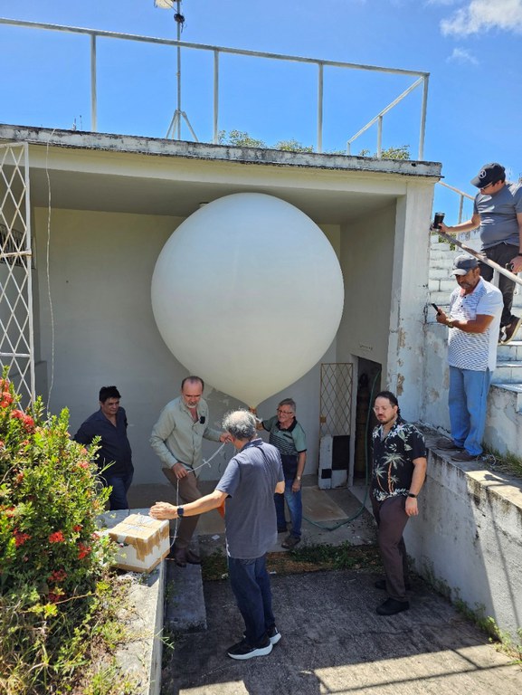 Equipe de seis pessoas realiza a preparação de um balão estratosférico branco antes do lançamento, em área aberta sob céu azul.