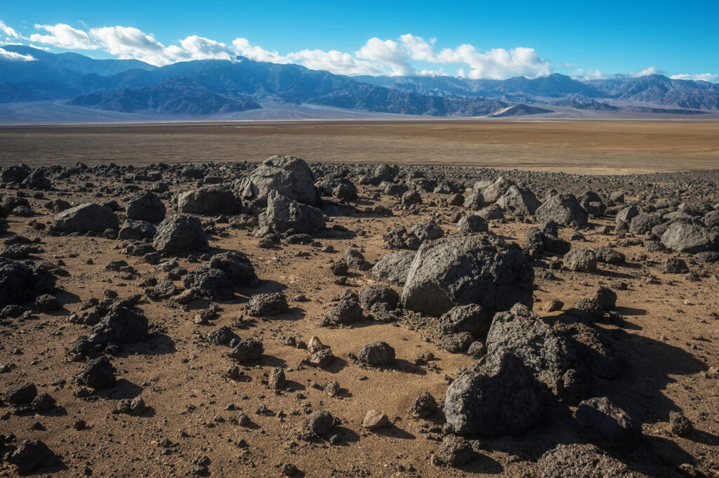 Rochas de basalto espalhadas sobre uma planície marrom em Mars Hill, no Death Valley National Park, com montanhas ao fundo e céu azul.