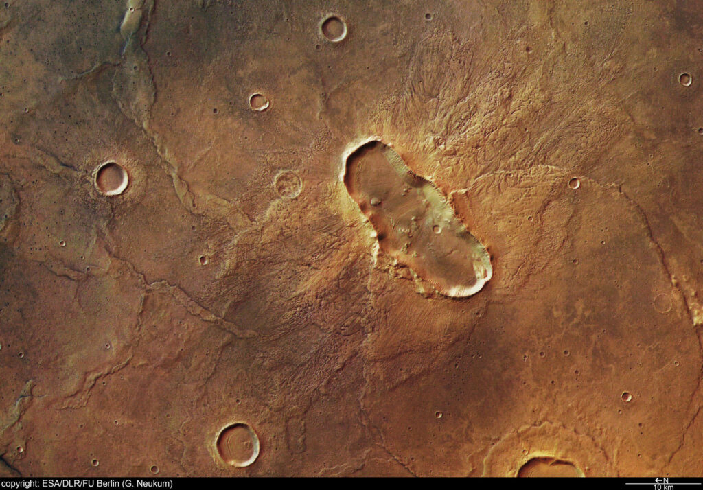 Imagem colorida da região de Hesperia Planum em Marte, mostrando uma grande cratera de impacto em formato de borboleta. A cratera elíptica apresenta bordas irregulares, interior sombreado e extensão alongada, destacando-se sobre a planície marciana amarelada e avermelhada.