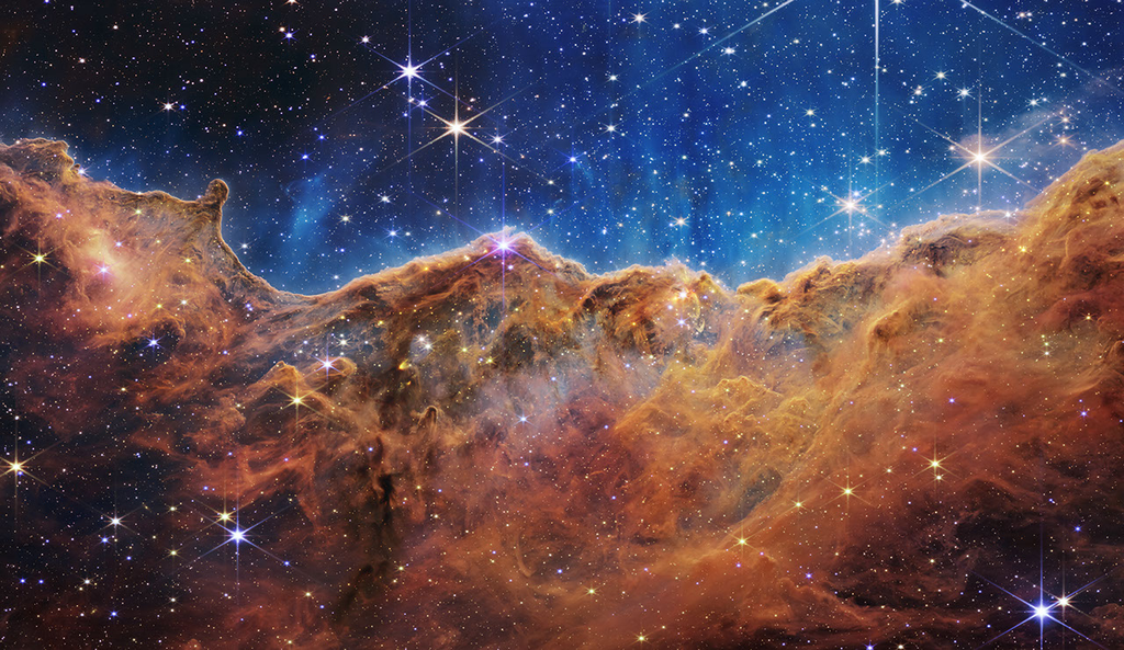 Imagem da Nebulosa de Carina (NGC 3324) capturada pelo Telescópio Espacial James Webb com a câmera NIRCam, mostrando nuvens de gás e poeira iluminadas em cores falsas do infravermelho próximo, com setas indicando norte e leste, barra de escala em anos-luz e legenda das cores dos filtros utilizados.