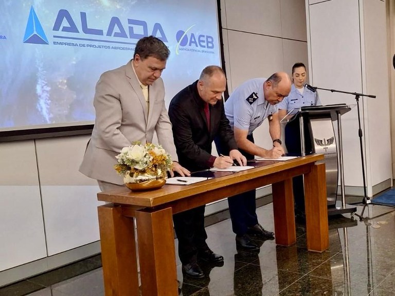 Cerimônia oficial com representantes da AEB, ALADA e COMAER assinando um Acordo de Cooperação Técnica para fortalecer o programa espacial brasileiro, com bandeiras e mesa de assinatura ao fundo.