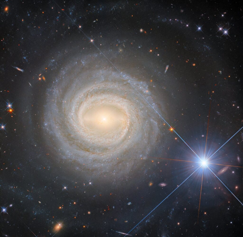 Imagem do telescópio Hubble mostrando a galáxia espiral barrada NGC 3783 vista de frente, com núcleo brilhante e braços espirais estreitos. À direita, uma estrela extremamente brilhante com picos de difração domina parte do quadro. Galáxias distantes e estrelas menores aparecem ao fundo.