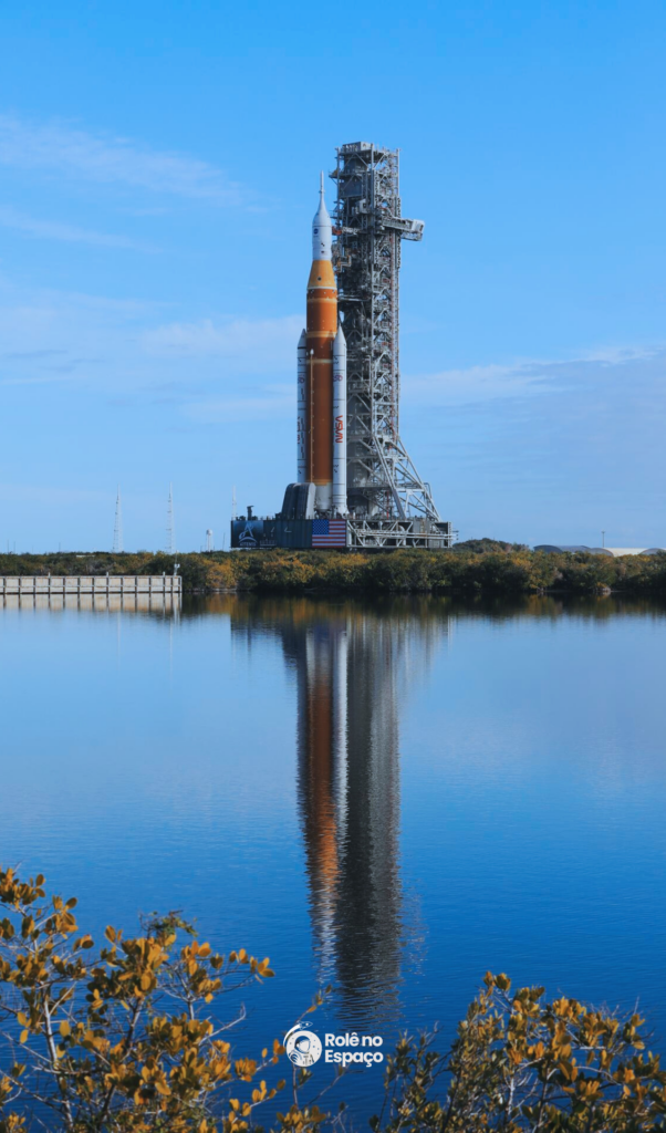 Foguete SLS da NASA refletido em um lago durante o rollout para a missão Artemis II, mostrando a estrutura completa e a plataforma de lançamento. Crédito: NASA.