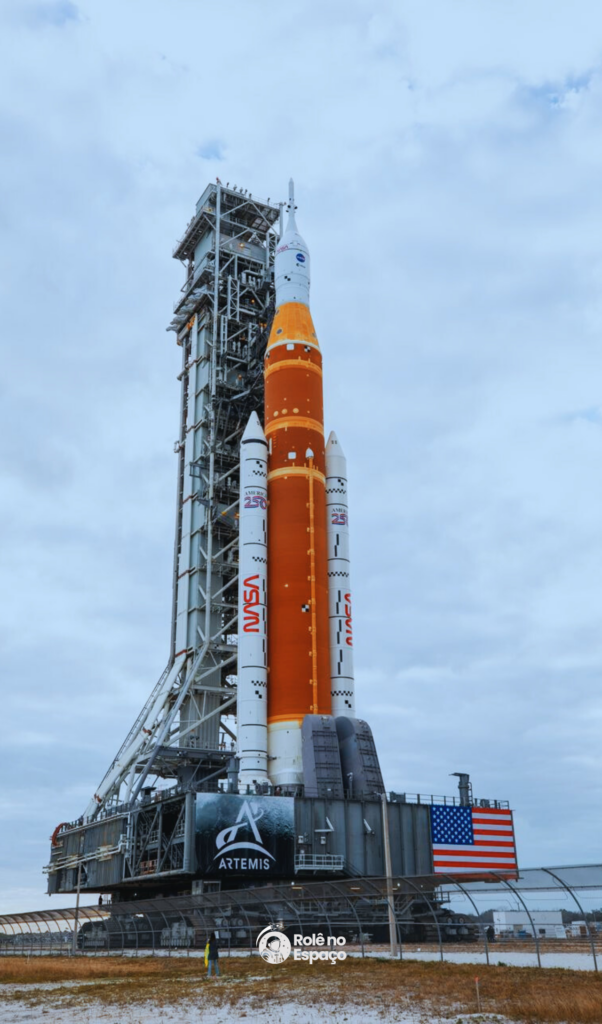 Foguete SLS da missão Artemis II em posição vertical na plataforma de lançamento do Kennedy Space Center, com torre de serviço ao lado e céu azul ao fundo. Crédito: NASA.
