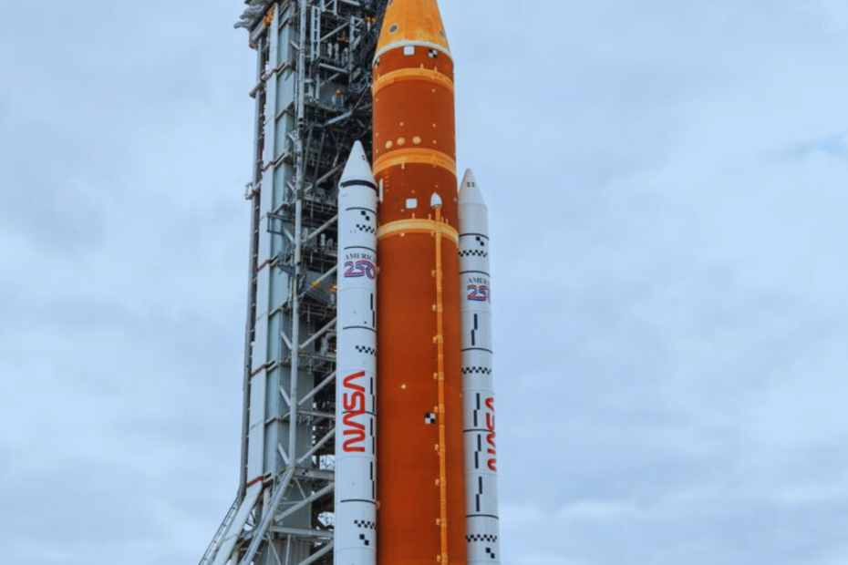 Foguete SLS da missão Artemis II em posição vertical na plataforma de lançamento do Kennedy Space Center, com torre de serviço ao lado e céu azul ao fundo. Crédito: NASA.