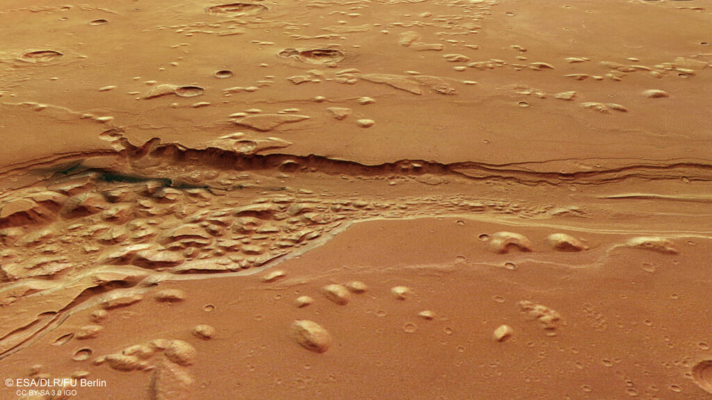 Vista aérea de Shalbatana Vallis em Marte mostrando um grande cânion sinuoso cercado por terreno irregular e superfícies suaves na região equatorial do planeta.