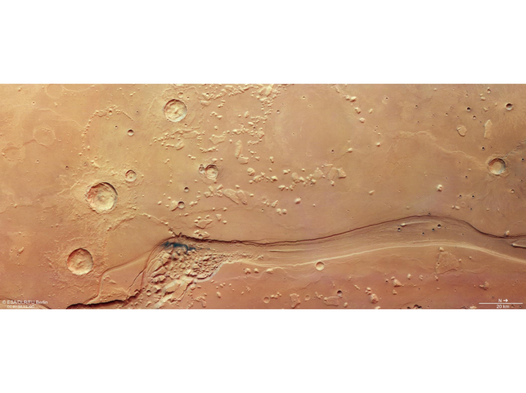 Imagem aérea de Shalbatana Vallis em Marte mostrando um grande canal sinuoso cercado por crateras e terreno caótico, registrado pela sonda Mars Express da ESA.